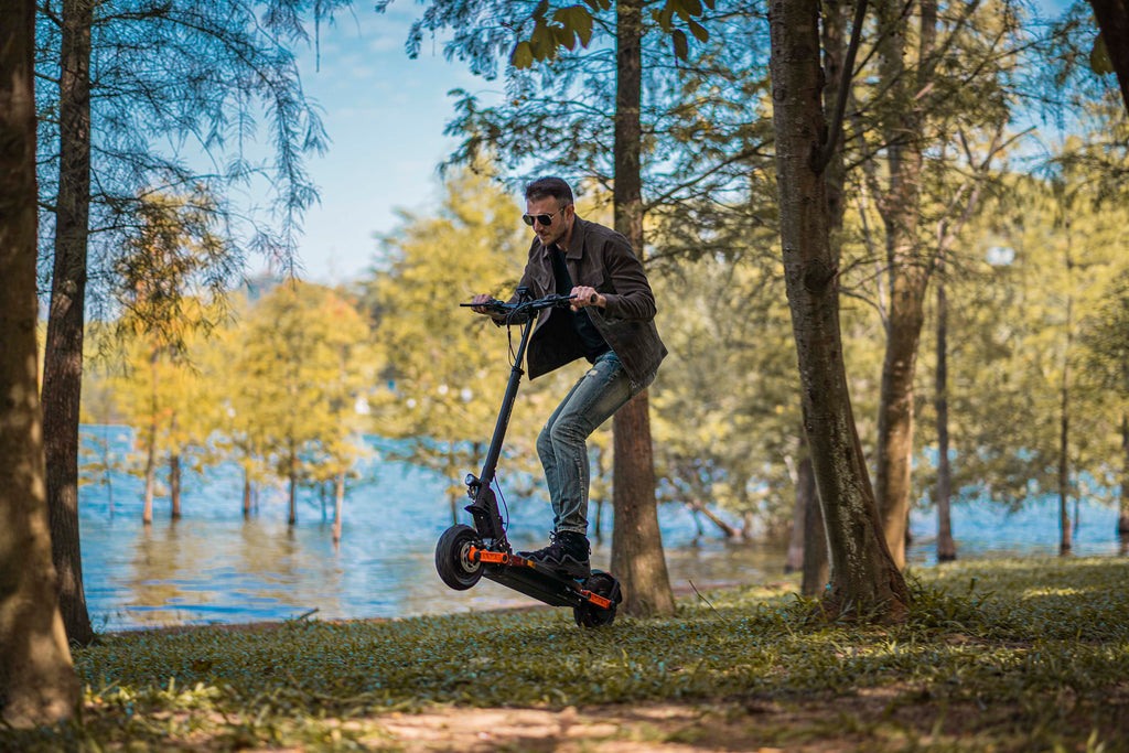 A man riding an Joyor S5Z electric scooter on a grassy path between trees near a lake, wearing a dark jacket, jeans, and sunglasses.