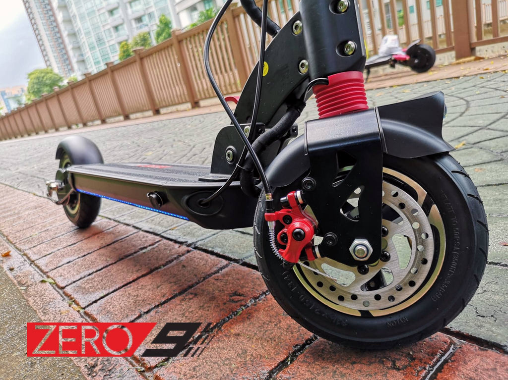 Close-up of a black electric scooter on a brick-paved outdoor walkway, showing the front wheel, disc brake, red front suspension, and part of the deck. A red and black 'ZERO 9' logo is visible in the lower left corner.