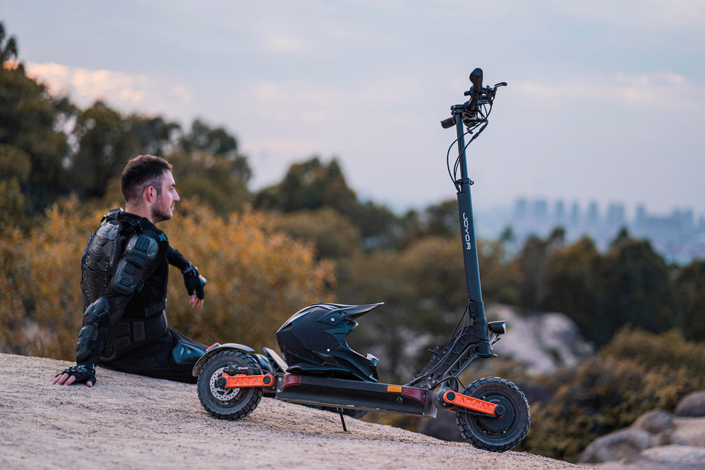 A man in black protective gear sits on a rocky outcrop next to a black Joyor S5Z electric scooter with orange accents, with trees and a distant city skyline in the background.