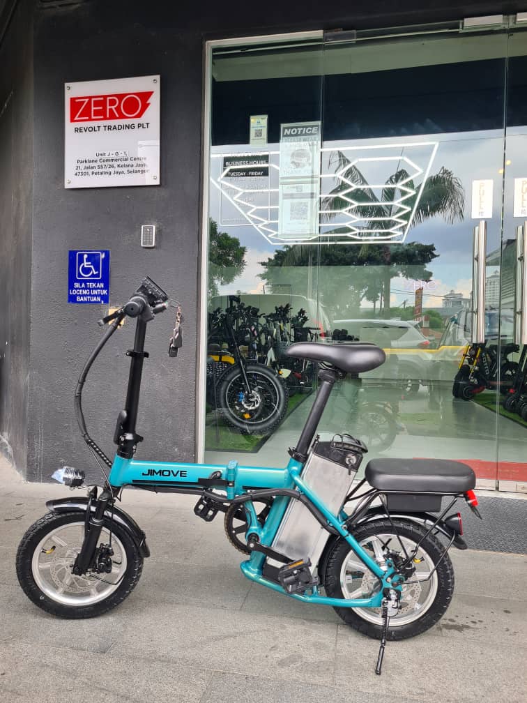 A turquoise and black Juke folding Jimove CC electric bicycle parked in front of a storefront with a glass door and a visible REVOLT Trading Ptd sign on the wall.