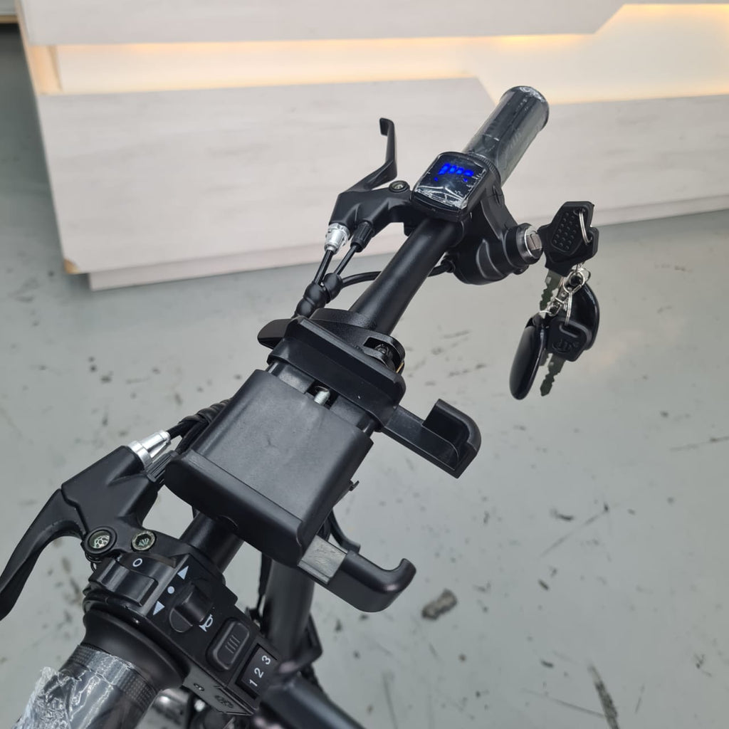 Close-up of a black Jimove CC folding electric bicycle handlebar setup with a mounted battery, digital display, brake levers, and keys, photographed indoors on a gray floor.
