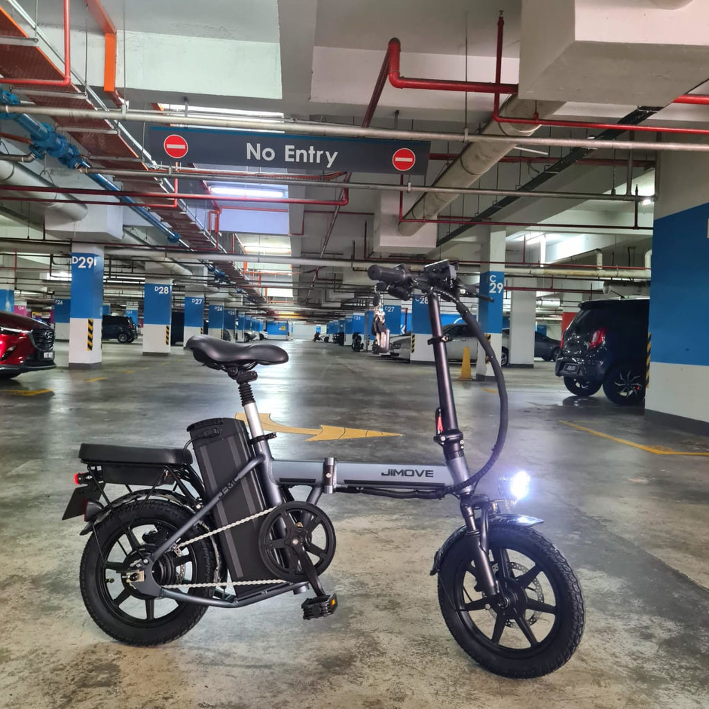 A black JINOVE CC folding electric bicycle with a rear battery pack, parked in a multi-level parking garage near Revolt Malaysia 