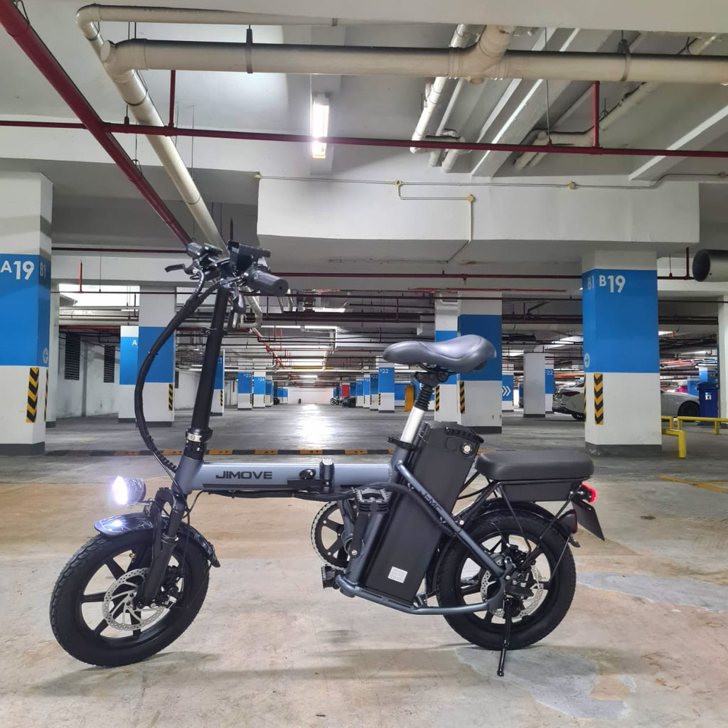 A dark gray folding Jimove CC electric bicycle with a rear storage bag is parked on its kickstand in a concrete and blue-and-white painted parking structure.