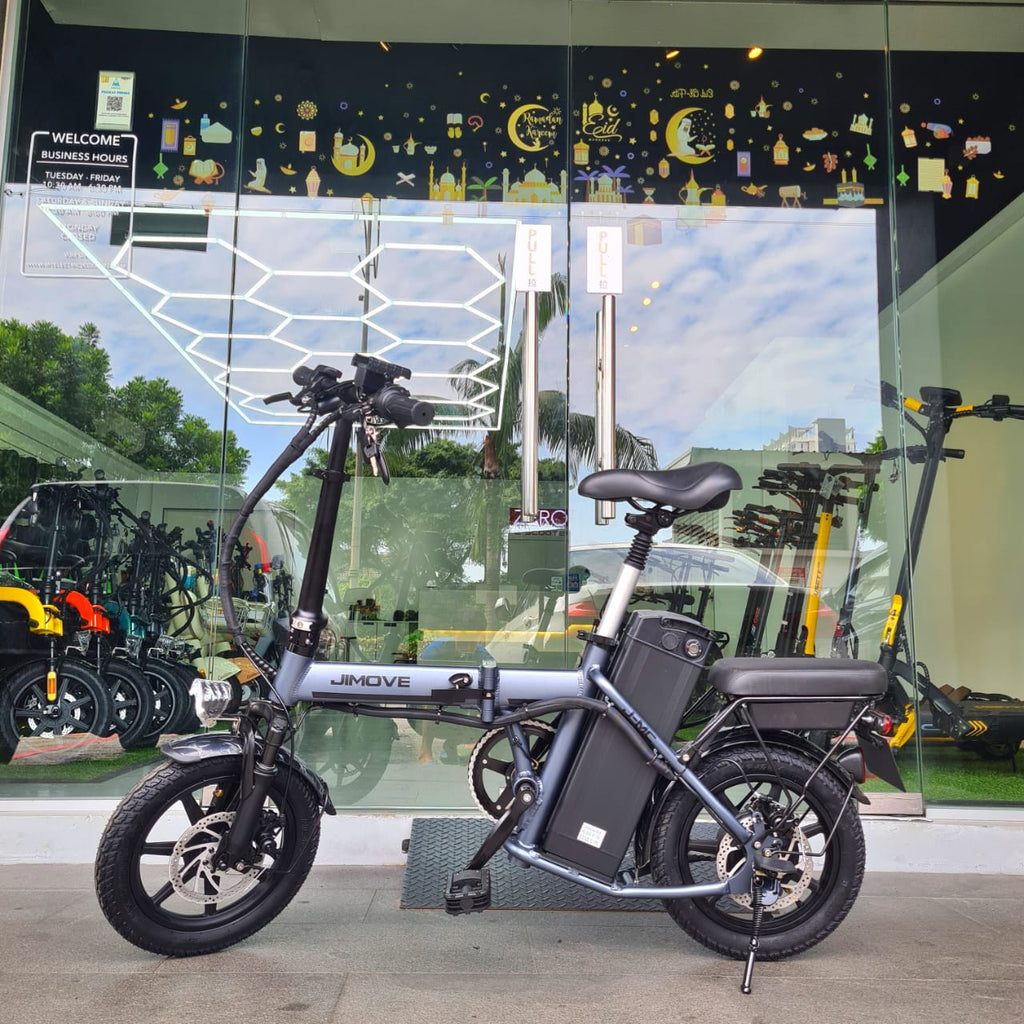 A JimoVE CC folding electric bicycle with a gray frame and black components, parked in front of a glass storefront window at Revolt Showroom PJ