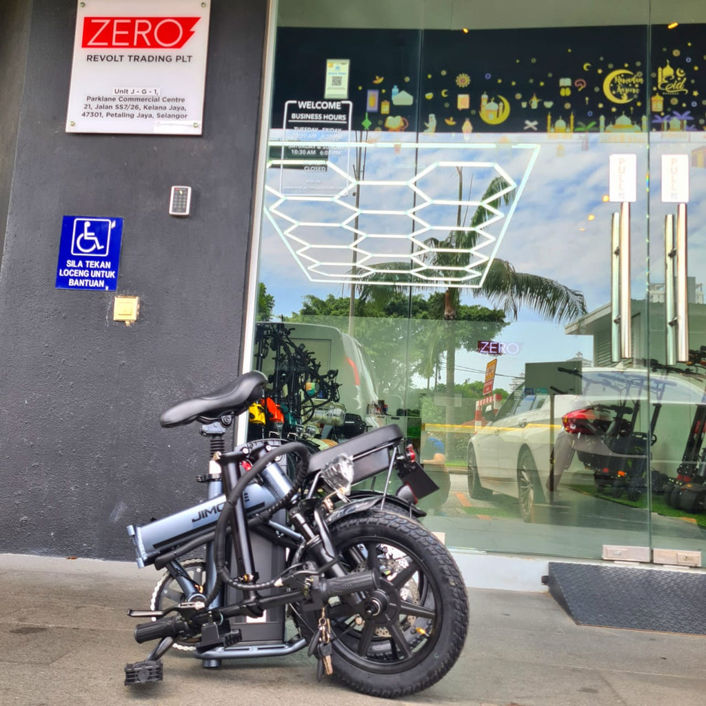 A folding Jimove CC electric bicycle with a light blue-gray frame and black components is parked in front of a glass storefront. The building has a dark gray wall with a red and white 'ZERO' sign and a blue handicap parking sign at Revolt Malaysia 