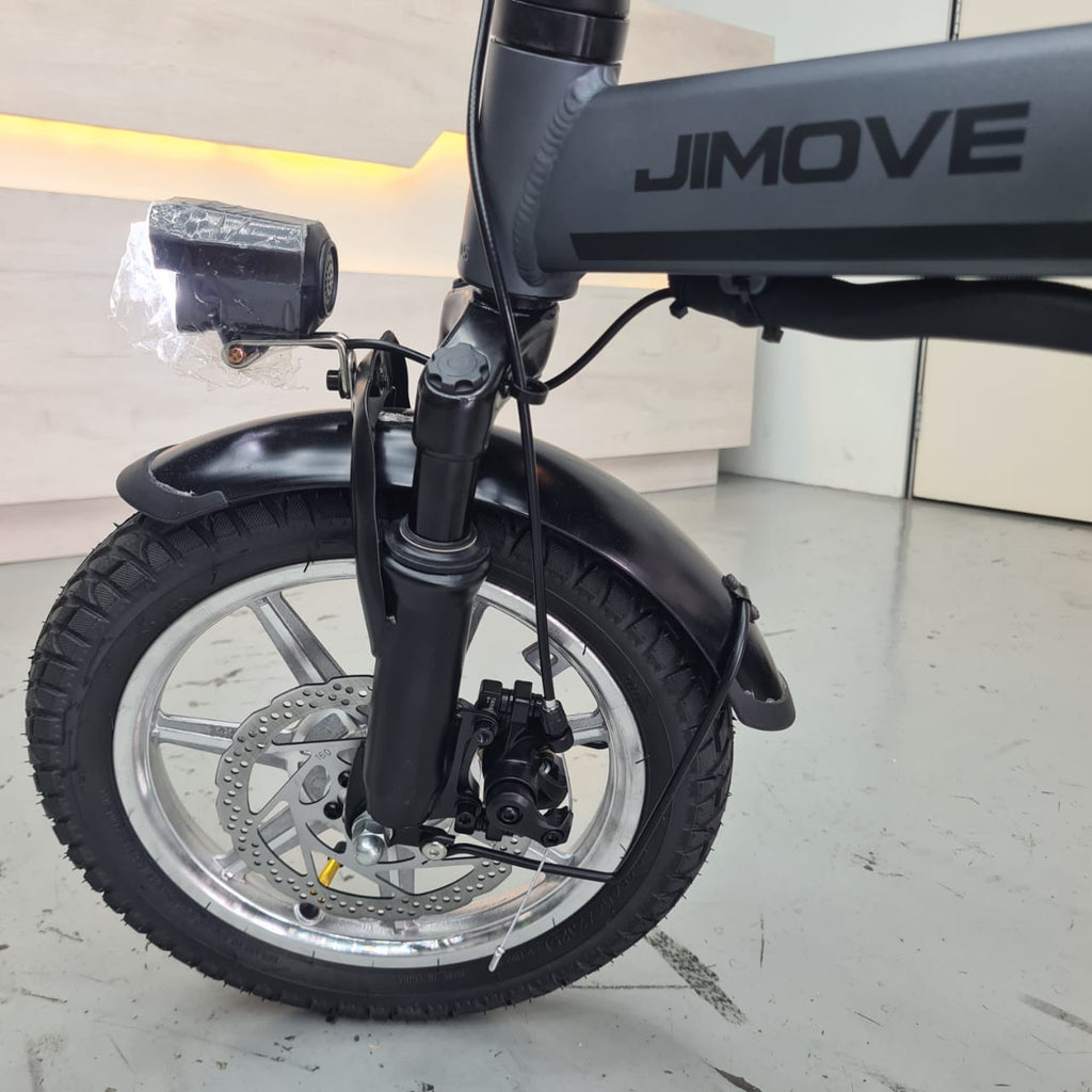 Close-up of the front wheel and fork of a JIMOVE CC folding electric bicycle, showing a black tire, silver wheel, disc brake, and black fender in an indoor showroom setting.