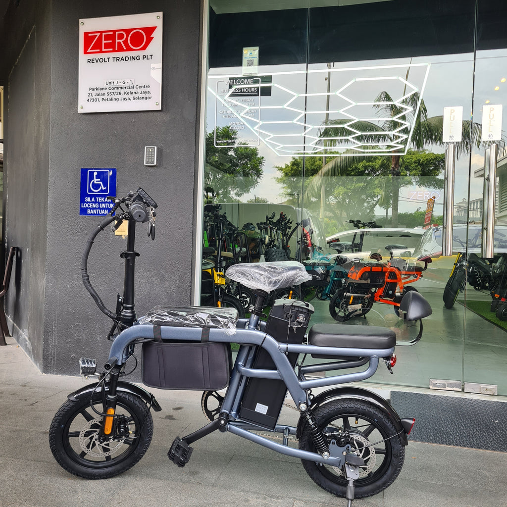 A silver Jimove M2 Pro electric bicycle with a rear step and black tires, parked on a sidewalk in front of a building with a glass window reflecting other scooters. A blue handicap parking sign is visible on the wall below.