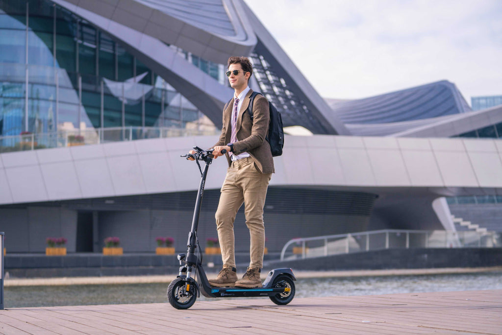 A man in a brown blazer, beige trousers, and sunglasses stands on a black Joyor C10 Removable Battery electric scooter on a waterfront promenade, with a modern glass building in the background.