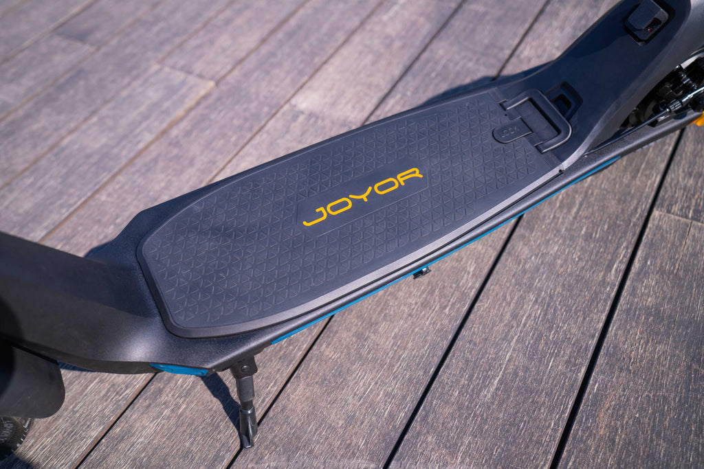 Close-up of a Joyor C10 Removable Battery electric scooter deck on a wooden deck, showing the textured black grip pad with yellow brand lettering.