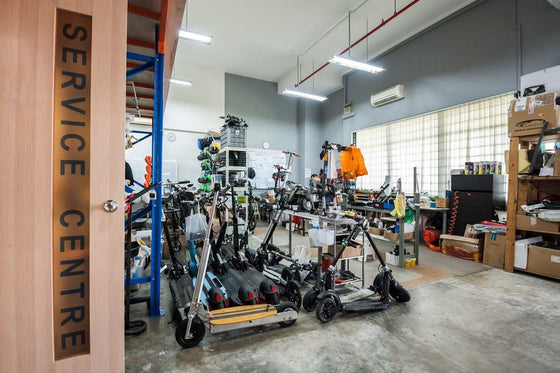 A large indoor service workshop with many electric scooters and bicycles displayed in a showroom area. A vertical wooden sign on the left reads 'SERVICE CENTRE'.