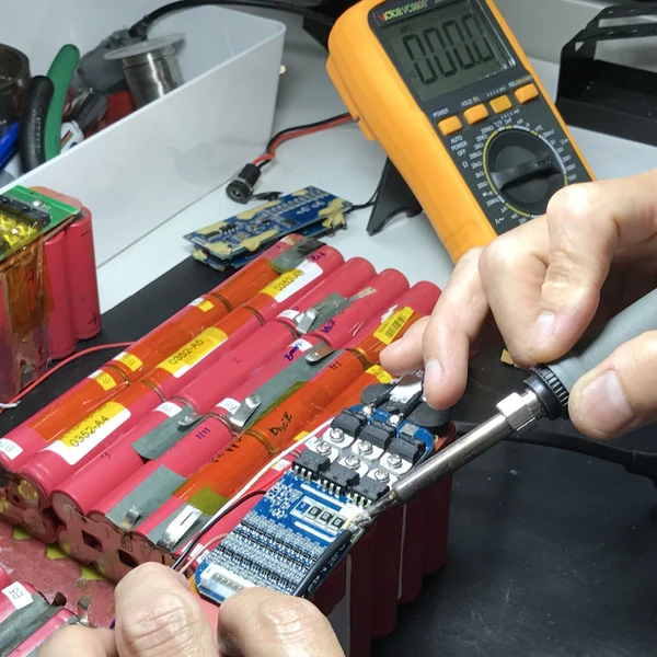 Hands soldering a small electronic circuit board with red cylindrical batteries in the foreground, with a multimeter and other electronics on a workbench.