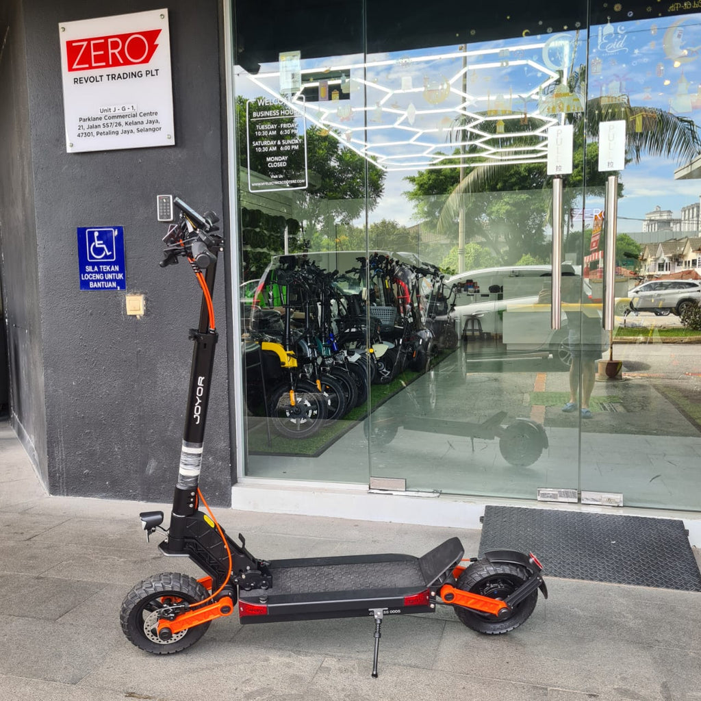 A black and orange JOYOR S5Z scooter is parked on a sidewalk in front of a glass storefront with a dark gray wall on the left. A blue handicap parking sign is mounted on the wall, and a white 'ZERO' Revolt Trading Plt sign is visible above it.