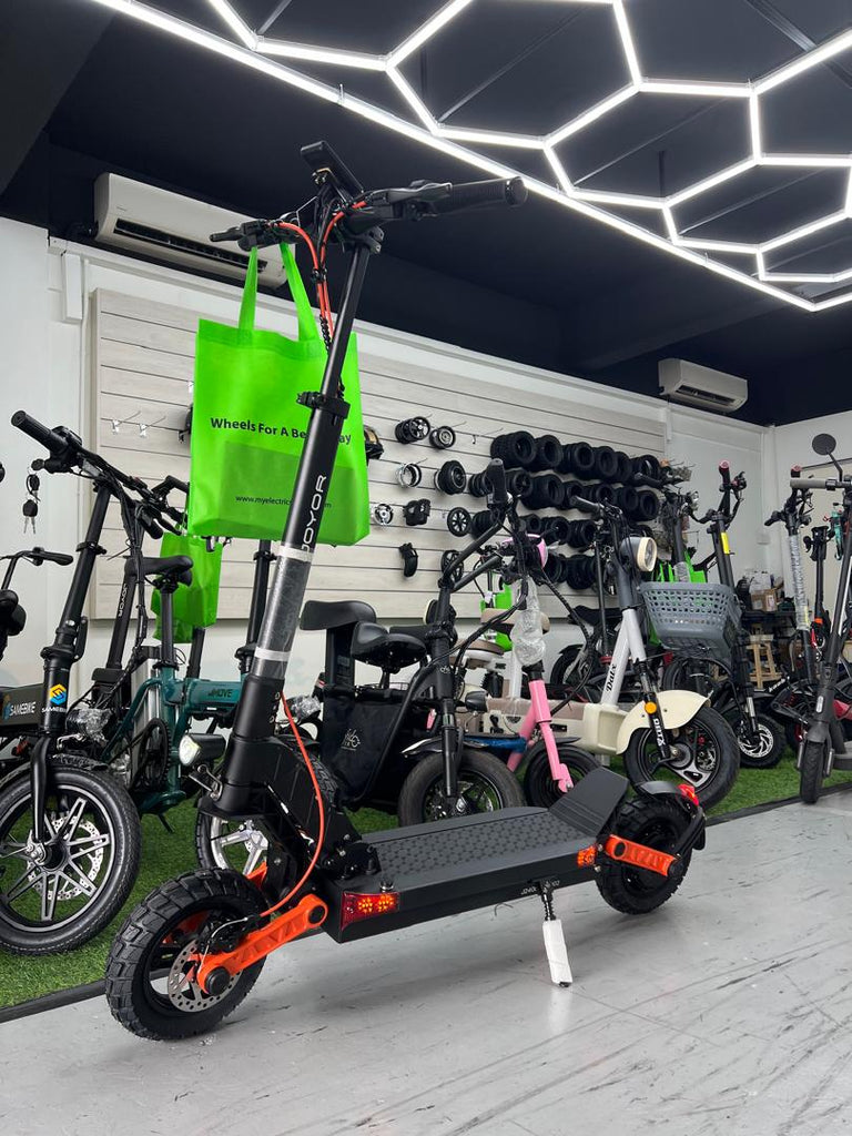 A black JOYOR S5Z FOLDING electric scooter with bright orange accents is displayed in a showroom with other electric bikes and a green tote bag hanging on the frame showroom display at Revolt Petaling Jaya.