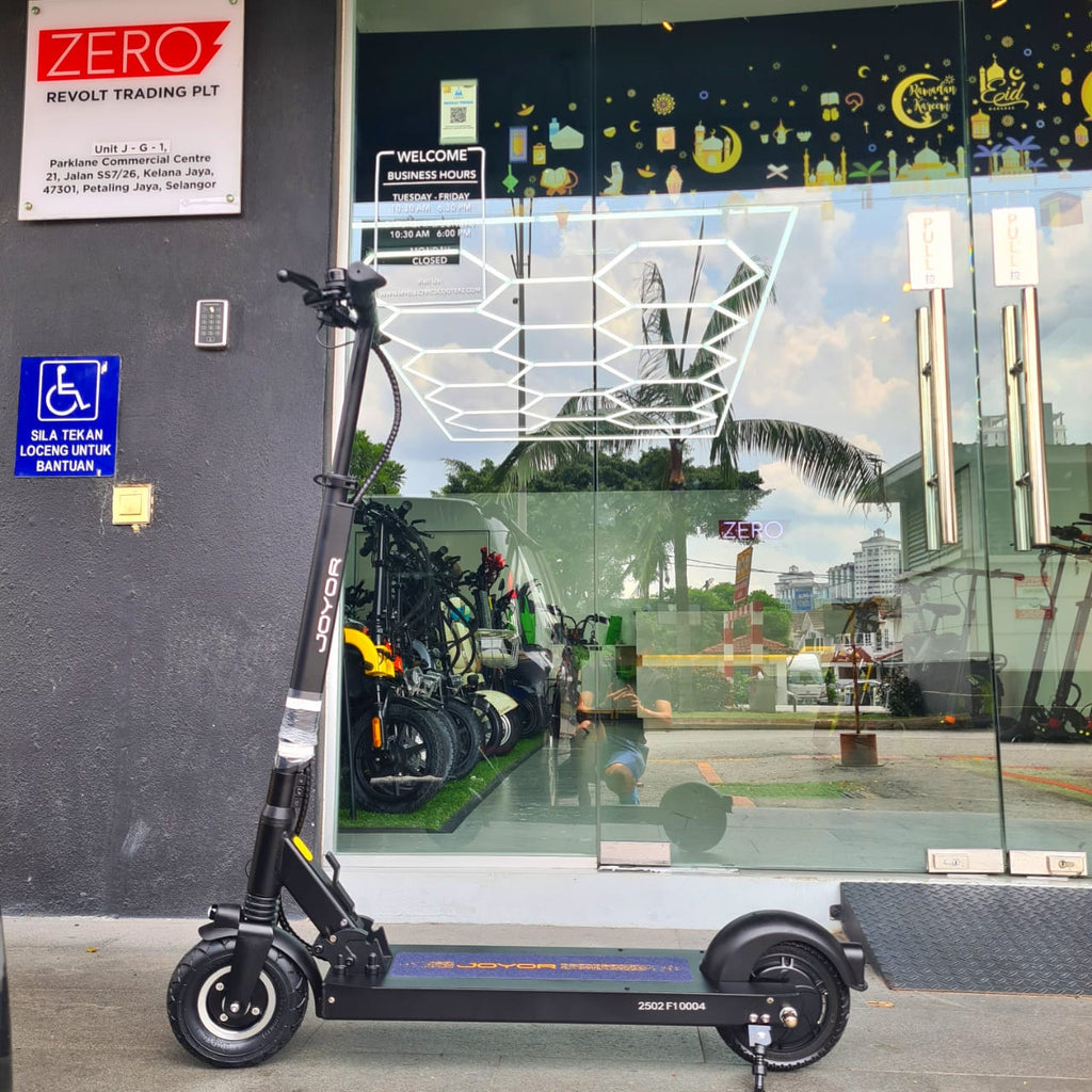 A black REVOLT joyor f1 electric scooter with a black frame and deck, parked in front of a glass storefront. The storefront has reflections of a tropical outdoor scene with palm trees and a person, and various shop signs are visible on the dark gray wall to the left.