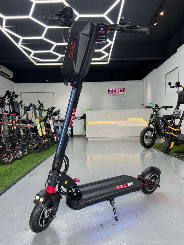 A black Zero 9 48V electric scooter with red and blue accents is displayed in a showroom. The scooter features a large pouch pack mounted on the handlebars, a step-through deck, and a kickstand, with the brand name 'ZERO' visible on the battery and deck.