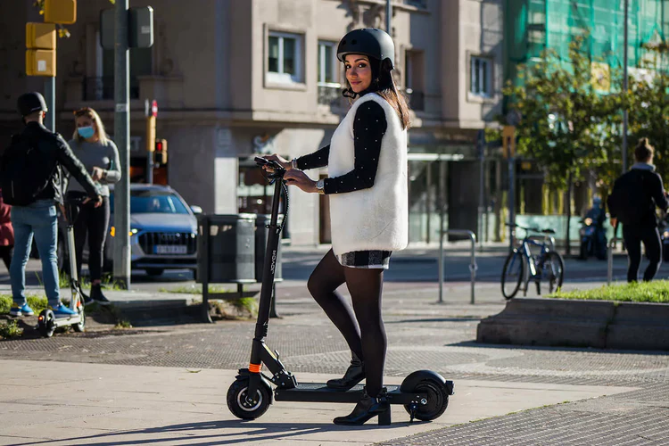 A woman wearing a black helmet and a white sleeveless vest over a black top rides a black joyor f1 lightweight foldable electric scooter on a city street.